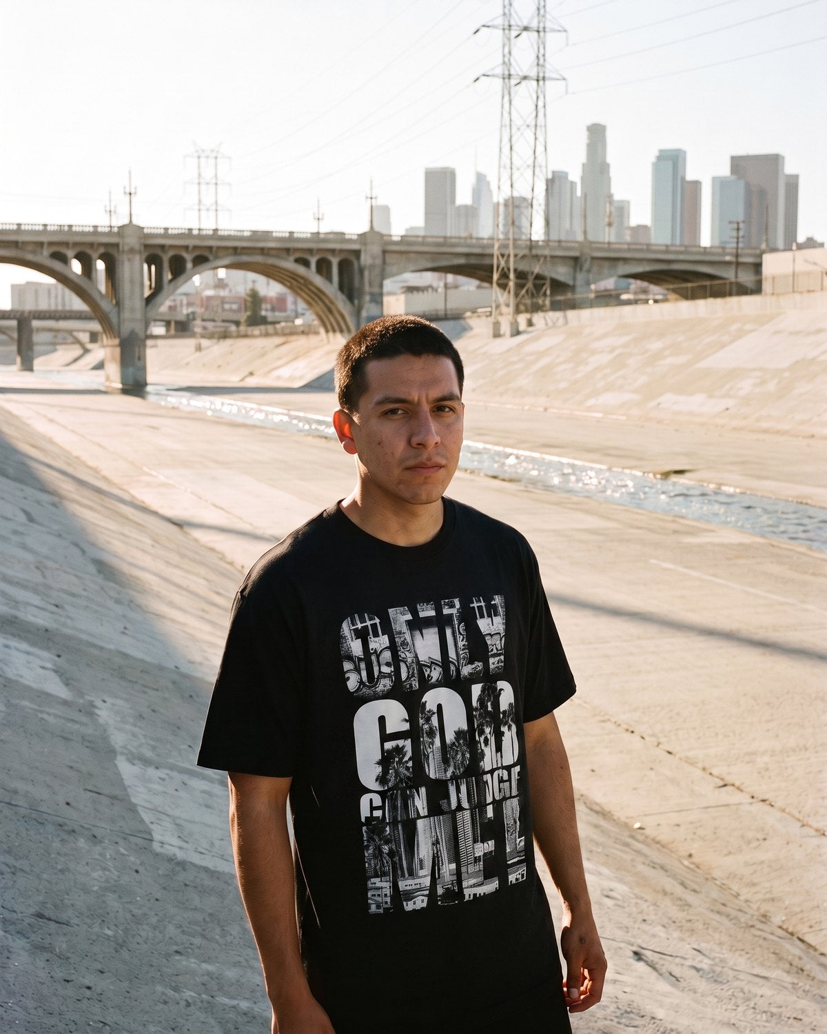 Only God Can Judge Me tee in the LA River channel, bridge and skyline behind.